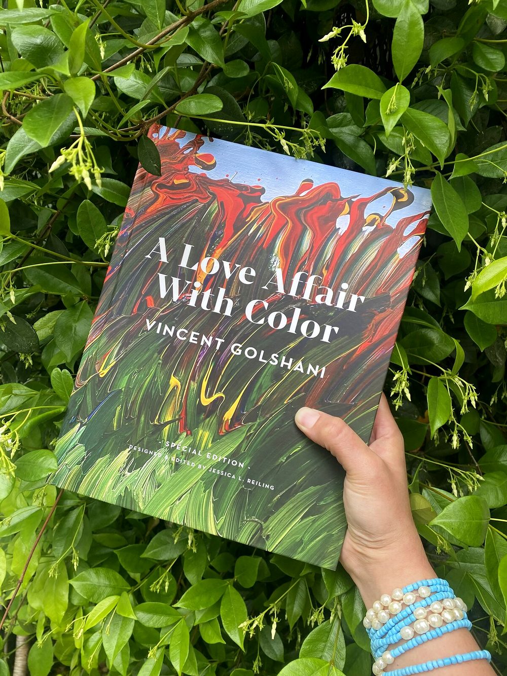A hand holding A Love Affair With Color by Vincent Golshani against green leaves